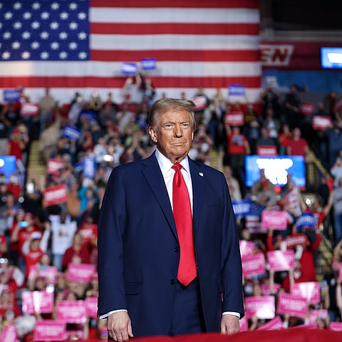 Donald Trump in a dark blue suit with a red tie stands in front of a large American flag. He is facing forward with a serious expression. Behind him, a large crowd is holding up red, white, and blue signs at what appears to be a political rally.