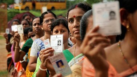 A medium-shot photo shows a long line of Indian women, many of them middle-aged, holding up their voter ID cards. The women are in a blurred outdoor setting with a yellow school bus visible in the background. They are wearing traditional Indian clothing in various colors and are waiting to cast their votes.