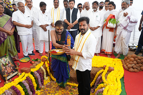 A photograph captures a traditional groundbreaking ceremony (Bhoomi Puja). The central figure, a man wearing a white shirt and a yellow and white scarf (Revanth Reddy, who is a prominent Indian politician), is standing in a partially dug pit adorned with yellow marigold garlands and surrounded by offerings like coconuts. He is looking down as a priest, who is kneeling and wearing traditional blue and saffron attire, is presenting a tray with colored powders (likely turmeric and kumkum) to him.
Behind them, a group of about a dozen men stand, mostly dressed in white traditional Indian attire (like dhoti and kurta), observing the ritual. One woman in a green saree is visible on the far left. The ground is covered with a red carpet. The ceremony appears to be a foundation-laying event for a new project.