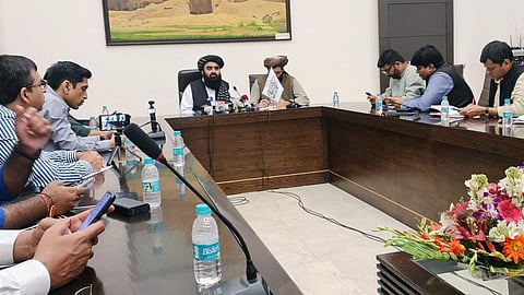 An image showing a press conference or meeting around a long, dark wood table. In the center, two men with beards wearing traditional clothing and turbans are seated. One of them, seated slightly to the right of the center, is holding a small white flag with what appears to be writing on it, possibly the flag of the Taliban (the Islamic Emirate of Afghanistan). Microphones are set up in front of them. Several other men, presumably journalists or attendees, wearing collared shirts and suits, are seated to the left and right sides of the table, many looking at or using their mobile phones. Several bottles of Bisleri water are on the table, along with a floral centerpiece with pink, orange, and white flowers on the right. In the background, there is a large framed picture depicting an ancient-looking structure.