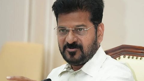 A head-and-shoulders, medium-close shot of Telangana Chief Minister Revanth Reddy, seated at a desk or table, speaking or making a point.
He is wearing a light-colored, possibly off-white, collared shirt. He has a dark beard and mustache, dark hair, and is wearing clear-framed rectangular glasses. He is looking slightly off-camera to the right with a serious expression and is extending his right arm forward, pointing with his index finger. A black microphone is visible just in front of him. The background is indoors and includes blurred details of light-colored furniture and a wall. A water bottle is visible on the right side of the desk.