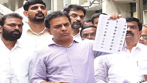 A medium close-up photo shows K. T. Rama Rao (KTR), dressed in a striped light purple shirt, standing amongst a group of men, all of whom appear to be politicians or party workers.
KTR is holding up a sheet of voter rolls, which he is presenting to the camera. His expression is serious. The other men are dressed in white shirts and look on intently. The background is an out-of-focus outdoor scene, possibly near a building.