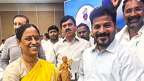 A full-length photo showing Konda Surekha, a woman in a bright yellow saree with a golden border and a patterned blouse, presenting a wooden figurine of a woman holding a bowl to Ponguleti Revanth Reddy, a man wearing a light blue collared shirt and light gray trousers.
Both Surekha and Revanth Reddy are smiling at the camera. They are standing indoors on a green carpet at what appears to be an official event. Several other men in formal or semi-formal attire are visible in the background, with one man's face partially cropped on the right. A banner with a blue and white design and the number "1" is visible behind them.