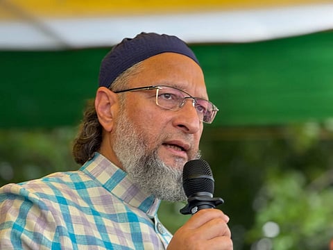 An eye-level close-up shot of Asaduddin Owaisi, an Indian politician, speaking into a microphone.
He is wearing a navy blue skull cap and a light blue, purple, and white checkered kurta. He has a gray beard and is wearing clear-framed eyeglasses. The background is blurred but shows green and white fabric, suggesting an outdoor political rally or press conference.