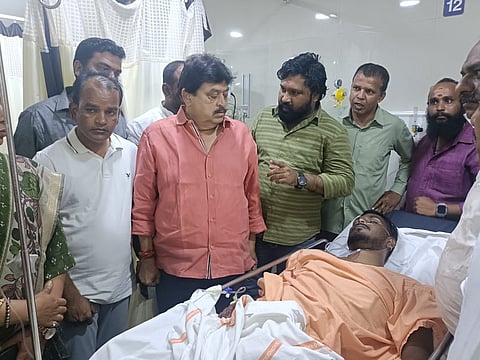 A group of men stand around a man lying on a hospital bed. The man on the bed is wearing an orange garment and has medical tubing attached to him. A prominent man in the center, wearing a salmon-colored shirt, is looking down at the injured man. Other men are gathered around them, some looking concerned. The setting appears to be a hospital room.
The image is a photo of BJP leader Ramchander Rao (in the salmon-colored shirt) and others visiting an injured man, Gau Rakshak Sonu Singh (lying on the hospital bed), in a hospital.