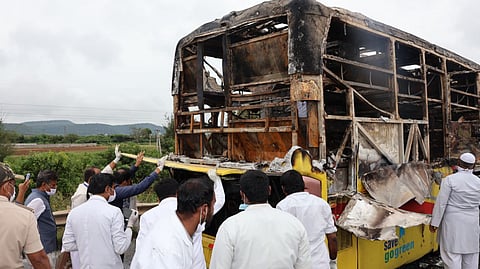 An image shows the burnt-out shell of a yellow-and-black bus following an accident. The rear section of the bus is completely destroyed by fire, with only the skeletal frame remaining.
Several men are gathered around the wreckage. Most are wearing white shirts or lab coats, and some are wearing masks and gloves, possibly first responders or officials investigating the scene. One man in a police uniform is visible on the far left. The ground appears to be a field or roadside area, with green foliage and hills in the distance under an overcast sky. A partially visible slogan on the lower side of the bus reads "save go green".
