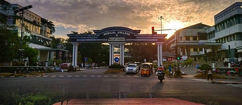 Entrance gate of Thiruvananthapuram Medical College at sunrise, with vehicles and people passing through the road in front of the campus.