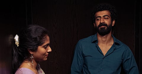 A woman and a man stand in a dimly lit space, facing slightly away from each other. The woman looks down with a faint smile, while the man gazes at her with a thoughtful expression. The background is dark wood paneling, adding to the intimate, introspective mood of the scene.