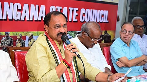 A political leader, Mahesh Kumar Goud, wearing a light yellow kurta and a traditional scarf with black and red patterns, is seated at a table and speaking into a microphone. He is holding a piece of paper in his left hand. Behind him, a banner with red and white text partially reads "TELANGANA STATE COUNCIL". Several older men are seated next to and behind him, listening. The man immediately to his right is wearing a white shirt and looking down, and the man further right is wearing a light blue shirt and glasses and looking forward.