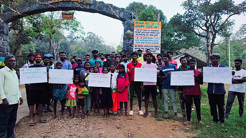 About 30 men, women and children holding placards in front of the Nanachi gate of the Nagarahole Tiger Reserve. Behind the group is the arch of the reserve. On either side of the the people are trees under a cloudy sky.