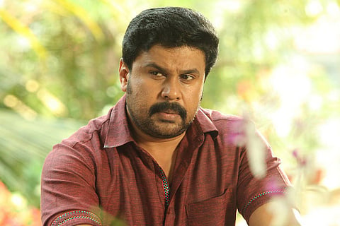A medium-shot portrait of actor Dileep, wearing a maroon or dark red collared shirt. He has a beard and mustache, and is looking slightly off-camera to the left with a serious or intense expression. The background is softly focused and appears to be an outdoor setting with green foliage and some lighter areas suggesting bright sunlight.