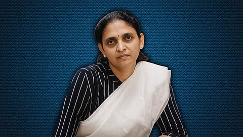 Portrait of a Judge Honey M Varghese wearing a white saree with a dark, striped blouse, seated against a textured blue background. She looks directly at the camera with a calm, serious expression, her hair neatly pulled back.