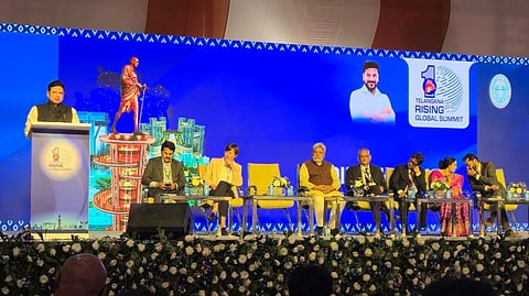 A low-angle photo captures a man speaking at a podium on a large stage, flanked by several dignitaries seated in a row.
The speaker, wearing a dark jacket over a white shirt, is to the left, standing behind a podium with the 'Telangana Rising Global Summit' logo. The backdrop is a large LED screen. On the screen, a graphic of a standing Mahatma Gandhi statue is visible to the speaker's left, and to the speaker's right, a portrait of a man (likely the Chief Minister or another prominent leader) is displayed next to the "Telangana Rising Global Summit" logo.
Seven people are seated on yellow chairs along the stage, facing the audience. They include men in both traditional Indian wear and suits, and one woman in a pink floral saree. The stage is decorated with white and purple floral arrangements along the front edge, and a large crowd is partially visible in the foreground.
