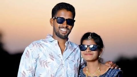 A medium shot of a young man and woman standing close together outdoors during sunset.
The man, on the left, has a dark complexion, a short beard, and is smiling. He is wearing a light blue button-down shirt with a subtle leaf pattern and dark sunglasses.
The woman, on the right, is wearing a traditional dark blue and silver lehenga with gold jewelry, including two necklaces, a waist belt, and a maang tikka on her forehead. She is also wearing dark sunglasses and has her arm resting behind the man.
The background is a soft, blurred golden-orange sky, suggesting dusk.
