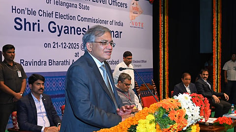 A medium shot of Shri Gyanesh Kumar, the Chief Election Commissioner of India, speaking at a podium during an event in Hyderabad on December 21, 2025.
He is a middle-aged man with graying hair and glasses, wearing a dark grey suit and a patterned blue tie. He stands behind a wooden podium heavily decorated with vibrant orange, yellow, and white marigold garlands.
In the background, a large white banner displays his name and title, along with the text "Ravindra Bharathi, Hyderabad." Several other officials in formal attire are seated on the stage behind him, and the setting is formal, featuring traditional floral decorations and green carpeting.