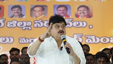 An image showing N. Ramchander Rao, a senior leader of the Bharatiya Janata Party (BJP) in Telangana, addressing a gathering. He is wearing a white shirt and holding a microphone in his left hand while gesturing with his right hand during a speech.
In the background, there is a large orange banner featuring Telugu text and portraits of other political figures. Several people are seated behind him, listening to the address. The setting appears to be a formal political event or meeting.
