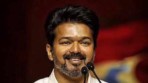 Vijay smiling from a podium. Two mics in front of him