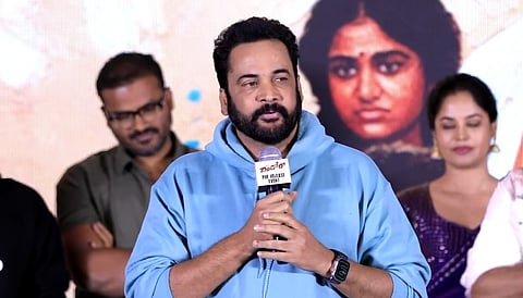 A medium shot of Telugu actor Sivaji speaking into a microphone at a pre-release event for the film Dhandoraa.
He is dressed in a light blue hooded sweatshirt and has a full black beard. He holds a microphone that has a white "Dhandoraa Pre-release Event" label on it. In the background, there is a large poster featuring a woman's face and several other people standing behind him, including a man in a green shirt and a woman in a purple saree.