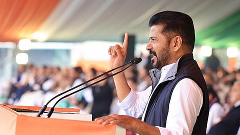 A medium shot from a side profile captures Revanth Reddy, the Chief Minister of Telangana, speaking passionately at an outdoor political rally. He is a middle-aged Indian man with short, dark hair and a well-groomed beard and mustache. He wears a crisp white long-sleeved shirt with the sleeves rolled up to his elbows, topped with a dark navy blue sleeveless quilted vest.
Standing behind a bright orange podium, he leans slightly forward with his left hand resting on the edge. His right arm is raised, with his index finger pointing upward in an assertive, emphatic gesture. Two microphones on long, thin stands are positioned in front of him.
The background shows a large, blurred crowd of people seated under a massive tent or canopy. The canopy features stripes of orange, white, and green, suggesting the colors of the Indian national flag. The lighting is bright and natural, indicating an afternoon event, creating a focused and energetic atmosphere.