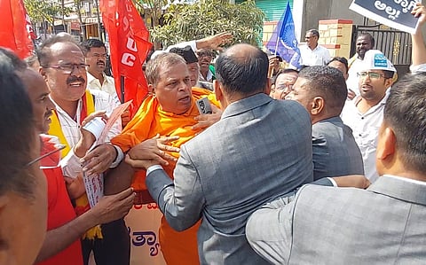 A group of protesters gathered on a street in daylight, with some holding flags and placards, as others appear to restrain or confront one protester during a demonstration.