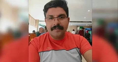 A medium shot of a middle-aged man with short dark hair, a mustache, and glasses, wearing a red and grey t-shirt. He is looking directly at the camera with a neutral expression. The background appears to be a brightly lit indoor space, possibly a restaurant or cafe, with other people seated at tables in the distance.