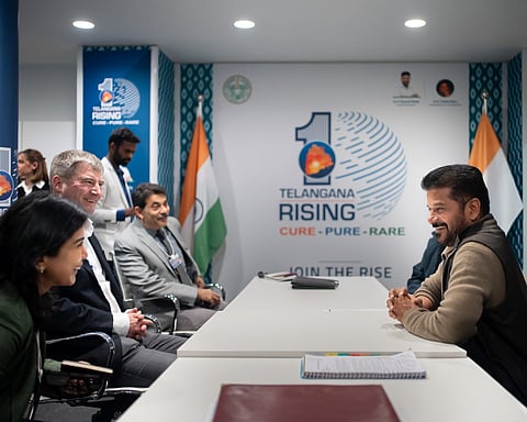 An indoor photograph shows a high-level meeting at the World Economic Forum in Davos. On the right, Revanth Reddy, the Chief Minister of Telangana, is seated at a white table, smiling and engaged in conversation. He is wearing a beige sweater over a dark vest.
Across from him on the left, three individuals are seated, including a woman in a green top in the foreground and a man in a dark suit. A man in a grey suit, Jayesh Ranjan, is also visible further down the table. Behind them stands an aide in a white blazer.
The background features a large "Telangana Rising" banner with the tagline "Cure - Pure - Rare" and the phrase "Join The Rise." The Indian national flag and a blue and white patterned pillar frame the setting. The overall atmosphere is professional yet warm and collaborative.