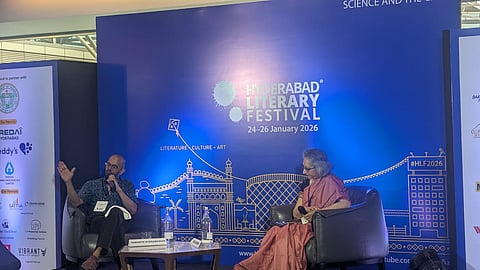 photograph of a stage at the Hyderabad Literary Festival 2026. Against a large blue backdrop featuring the festival logo and line art of Hyderabad landmarks, two speakers are seated in grey armchairs. On the left, Samanth Subramanian, a man with a beard and glasses, gestures with one hand while speaking into a microphone. On the right, Usha Raman, a woman in a pink and black saree, sits listening. A small wooden coffee table with water bottles and nameplates sits between them. Panels with sponsor logos are visible on either side of the stage.
