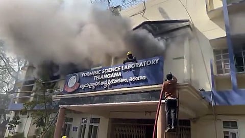 A wide, medium shot shows dark grey smoke billowing from the second-story windows of the Forensic Science Laboratory building in Telangana.
The building is a multi-story, cream-colored structure with blue trim. A large blue sign above the main entrance clearly identifies the facility in both English and Telugu. In the foreground, a firefighter wearing a yellow helmet is visible on a balcony near the smoke, while another person in a brown shirt climbs a ladder leaning against the building’s entrance overhang. The scene captures an active emergency response to a structural fire.