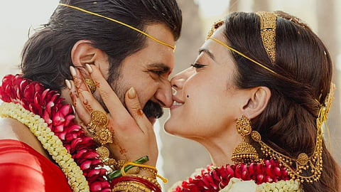 A close-up, intimate profile shot of a couple in traditional Indian wedding attire, leaning in close with their noses touching. The bride, on the right, wears an ornate gold-embroidered red and orange saree with heavy gold jewelry, including a maang tikka, jhumkas, and intricate armbands. Her hand gently cups the groom's face. The groom, on the left, wears a red garment and has a mustache and long hair. Both are adorned with thick garlands of red and white flowers. The lighting is soft and warm, highlighting their joyful expressions against a blurred natural background.