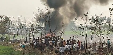 Gemini said
An outdoor scene shows a large crowd of people gathered in a rural, grassy area near several thin, leafless trees. Thick, dark grey smoke billows into the sky from behind the trees, where a small fire is visible. The crowd is composed of many people standing and looking toward the source of the smoke. In the background, there is a green field and a few palm trees under a hazy sky.
