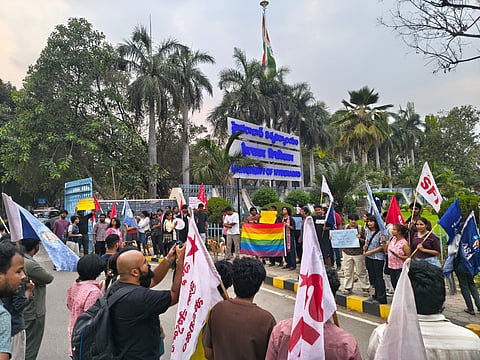 A group of students at the entrance of the University of Hyderabad (HCU) are gathered for a protest. Several students are holding flags from various student organizations, including the SFI (Students' Federation of India) and ASA (Ambedkar Students' Association). In the center, two students hold a large rainbow pride flag, while others carry handmade placards. The university's main gate and blue signage are visible in the background under a cloudy sky. The scene is active, with some individuals taking photos and others standing in a semi-circle.