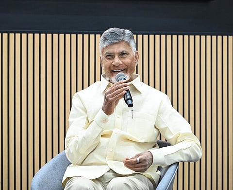 A medium shot of N. Chandrababu Naidu, the former Chief Minister of Andhra Pradesh, sitting in a modern grey chair. He is smiling warmly while holding a microphone to his mouth as if speaking at an event.
He is dressed in his signature light-cream, short-sleeved button-down shirt and matching trousers. The background features a minimalist wall with vertical light-wood slats over a dark panel. The lighting is bright and professional, highlighting his grey hair and expressive gestures.