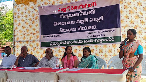 A wide-angle outdoor shot of a public hearing in Gabbilalapet, Telangana. Five people (three men and two women) are seated behind a long table covered with white and red cloths. To the right, a woman in a peach-colored floral saree stands, speaking into a microphone.
Behind the group is a large banner written in Telugu saying a Telangana Public School should be established in Gabbilalapet.
The setting is under a temporary tent structure with blue and white fabric overhead and a patterned yellow backdrop. The atmosphere appears formal and community-focused.