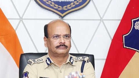 A formal portrait of B. Shivadhar Reddy, a high-ranking official of the Telangana Police, seated at a wooden desk with his arms crossed.
He is wearing a khaki police uniform featuring several service ribbons on his chest, a name tag, and shoulder insignia indicating his rank. He has short, thinning dark hair, a mustache, and wears gold-rimmed glasses and a gold watch.
In the background, a large Telangana Police emblem is mounted on a white geometric-patterned wall. The emblem includes the State Emblem of India and the motto "Duty, Honour, Compassion." To his left is the Indian National Flag, and to his right is the Telangana Police flag. A microphone, a bottle of water, and some tissues are visible on the desk in the foreground.
