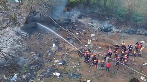 Firefighters in protective gear spray water over a charred, debris-strewn site of a fireworks unit explosion in Mundathikode near Thrissur, with smoke rising from burnt ground and scattered remains visible around them.