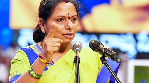 A medium shot of Kalvakuntla Kavitha, an Indian politician, speaking at a podium. She is wearing a lime green saree with a blue border and has her right hand raised with her index finger pointed upward as if making a strong point. Behind her, a large digital screen displays the letters "TRS" in white over a blue map of Telangana against a yellow background. The lighting is bright and stage-focused, emphasizing her animated expression.