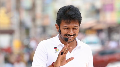 A medium shot shows Udhayanidhi Stalin, a smiling man with short dark hair and a light mustache, wearing a white polo shirt with a small emblem on the chest. He is waving his right hand toward the camera while wearing a black headset microphone. He stands behind a black podium or vehicle panel that features three black-and-white portraits of prominent Dravidian leaders: Periyar E. V. Ramasamy, C. N. Annadurai, and M. Karunanidhi. The background is a brightly lit, blurred outdoor urban setting.