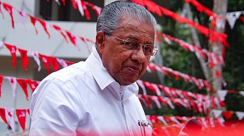Kerala CPI(M) leader Pinarayi Vijayan wearing a white shirt and glasses, standing outdoors during an election campaign event with red CPI(M) flags and buntings visible in the background.