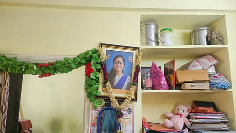 An indoor eye-level medium shot shows a framed portrait of a woman hanging on a pale yellow wall. The woman in the portrait is smiling, wearing glasses and a blue and white patterned saree, with a garland of yellow and purple flowers draped over the frame. A separate garland of green leaves and red hibiscus flowers hangs to the left of the frame. Below the portrait, there is a calendar and a small blue-framed mirror. To the right of the portrait, there are three built-in wall shelves containing various items including stainless steel containers, a pink stuffed bear, and a stack of books and papers. In the background, on the lower left, a portion of a kitchen is visible with a gas stove and several small containers on a shelf. The lighting is bright and even across the scene.