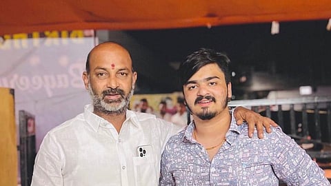 An eye-level medium shot captures two men standing side-by-side. The older man on the left, with a bald head and a graying beard, wears a white button-up shirt and has a red mark on his forehead. He rests his left arm around the shoulder of the younger man on the right, who has dark hair, a mustache, and a goatee. The younger man wears a light blue patterned button-up shirt and a black smartwatch on his left wrist. The background is blurred, showing a warm-toned canopy and some structures.