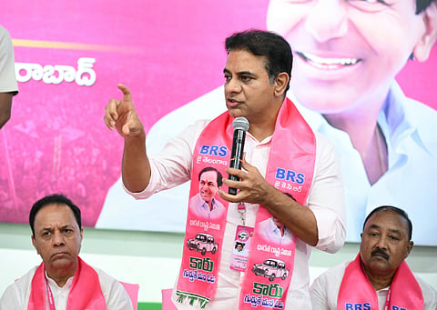 An indoor medium shot of K. T. Rama Rao (KTR), a prominent Indian politician from the Bharat Rashtra Samithi (BRS) party, speaking into a microphone during a political event. He is wearing a light-colored shirt and a bright pink party scarf draped around his neck. The scarf features the BRS logo, an image of a car, and a portrait of K. Chandrashekar Rao. KTR is gesturing with his right hand while holding the microphone in his left. Two other men, also wearing pink party scarves, are seated behind him. A large pink backdrop with a partially visible portrait of K. Chandrashekar Rao is behind them.