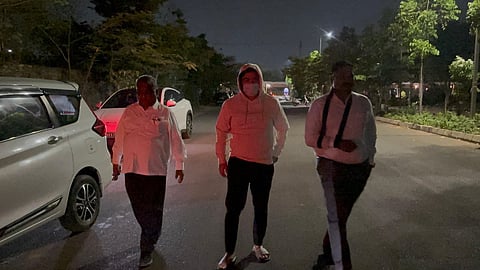 A low-angle, eye-level full shot shows three people walking towards the camera on a paved road at night. On the left, an older person wears a white long-sleeved button-down shirt and dark pants. In the center, a person wearing a white hoodie with the hood up, a white face mask, dark pants, and white sandals walks with their hands in their pockets. On the right, another person wears a white long-sleeved button-down shirt and dark pants. The road is dark, and the figures are illuminated by an off-camera light source. Several cars are parked on the left side of the road, and trees line both sides. In the background, streetlights and a building are visible under a dark sky.