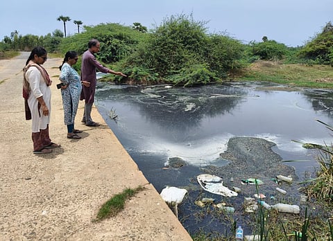 Three people stand on a concrete path, looking down at a heavily polluted body of water. The water is dark and murky, covered in patches of white foam and a thick, dark film. Plastic bottles and other trash float in the foreground near some tall grass. Behind the water, there are dense green bushes and trees under a clear sky. One person is pointing toward the middle of the water.