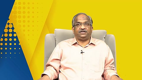A man with glasses and a light pink shirt sits in a grey chair against a bright yellow and blue background. The background features a geometric pattern with dots and diagonal lines. The man is looking directly at the camera with a neutral expression.