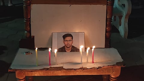 The image shows a somber memorial scene at night. A small wooden table, placed on a hexagonal-tiled ground, serves as an altar. Atop the table sits a framed portrait of a young man with dark hair and glasses. Several lit candles of various colors (yellow, pink, and white) are arranged in front of the portrait and on the ground surrounding the table, their flames casting a warm glow in the dark environment. In the background, blurred lights and a park bench with graffiti are visible.