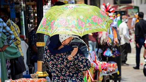 A woman in a floral print dress carries a sleeping baby on her shoulder while walking through a sunny outdoor market in Amritsar, India. She holds a bright lime-green umbrella decorated with pink roses to shield the child from the sun. The baby wears a white bandana with a religious symbol and denim overalls. In the background, vendors display colorful bangles and merchandise under the bright daylight.