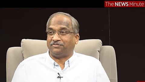 A middle-aged man with salt-and-pepper hair, a mustache, and glasses is seated in a light-colored armchair against a dark background. He is wearing a light blue button-down shirt with a small black lapel microphone attached. In the top right corner, there is a logo for "The NEWS Minute."