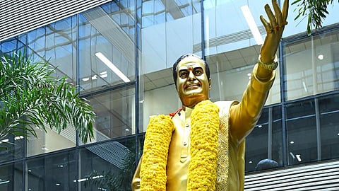 A low-angle, full-length shot shows N. Chandrababu Naidu, a man with grey hair, dressed in a yellow shirt and light-colored trousers, standing with his hands folded in prayer before a large, golden statue of N.T. Rama Rao. The statue, depicting the legendary actor and politician with one arm raised in a wave, is adorned with a thick garland of yellow marigolds. The base of the statue is also heavily decorated with layers of orange and yellow marigold garlands and several bouquets of colorful flowers, including white, purple, and yellow blossoms. The scene takes place outdoors, against the backdrop of a modern building with large glass windows. To the left, a few green plants are visible, adding a touch of nature to the setting.