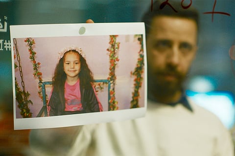 A still from The Voice of Hind Rajab showing a photograph of a young girl pinned on a glass surface, with a man standing blurred in the background.