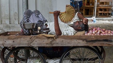 A side-view, medium shot shows a male street vendor lying down to rest on his wooden cart. He wears a light-colored, short-sleeved shirt and a grey plaid lungi. He rests his head on a small brown burlap sack next to a pile of red onions and holds a traditional fan made from dried palm leaves above his face with his right hand. A metal weighing scale sits near his feet. The cart has large, wire-spoked bicycle wheels.